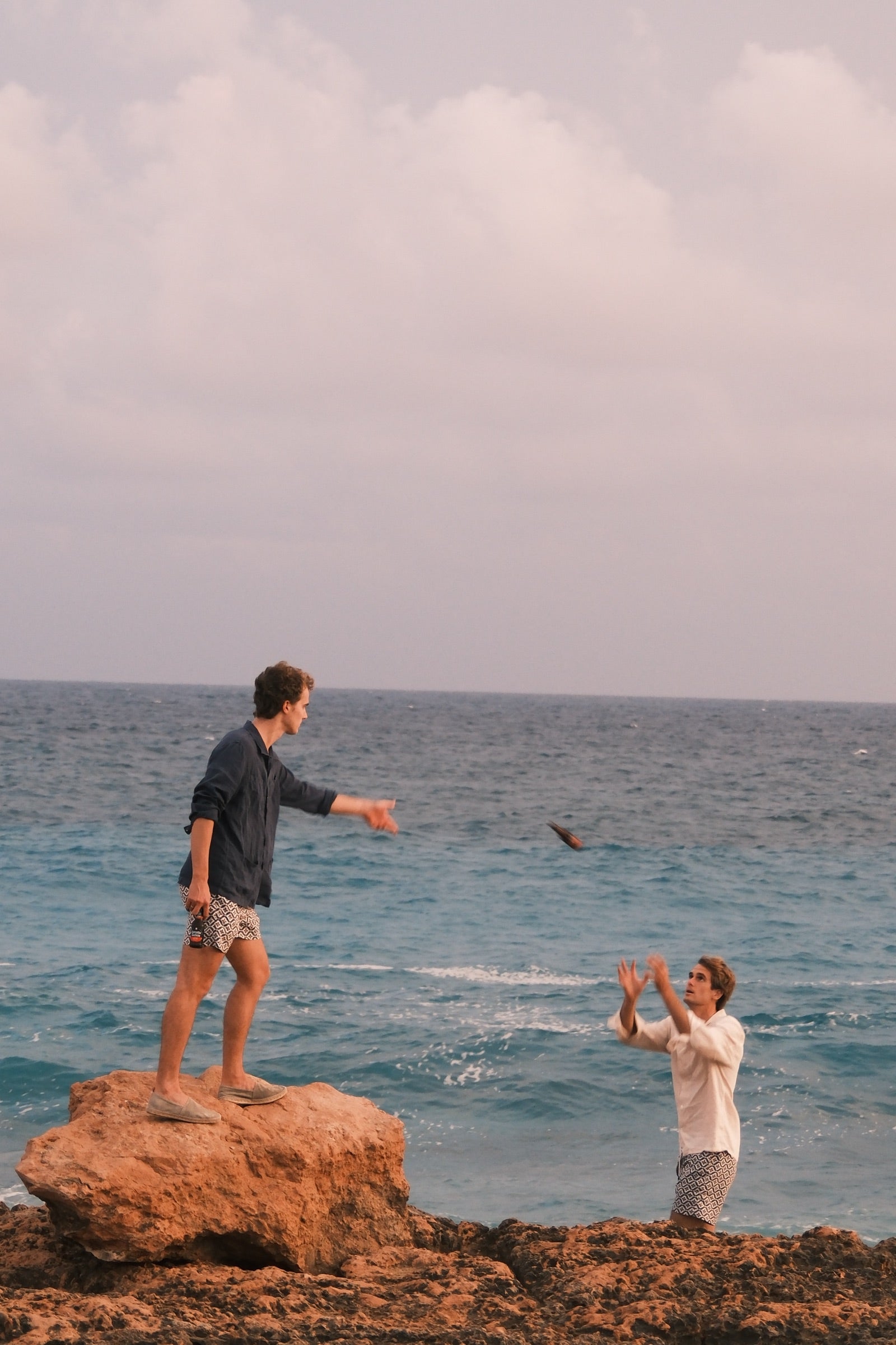 Dos jóvenes junto al mar turquesa, uno de pie sobre una roca lanzando un objeto y el otro con los brazos alzados para cogerlo