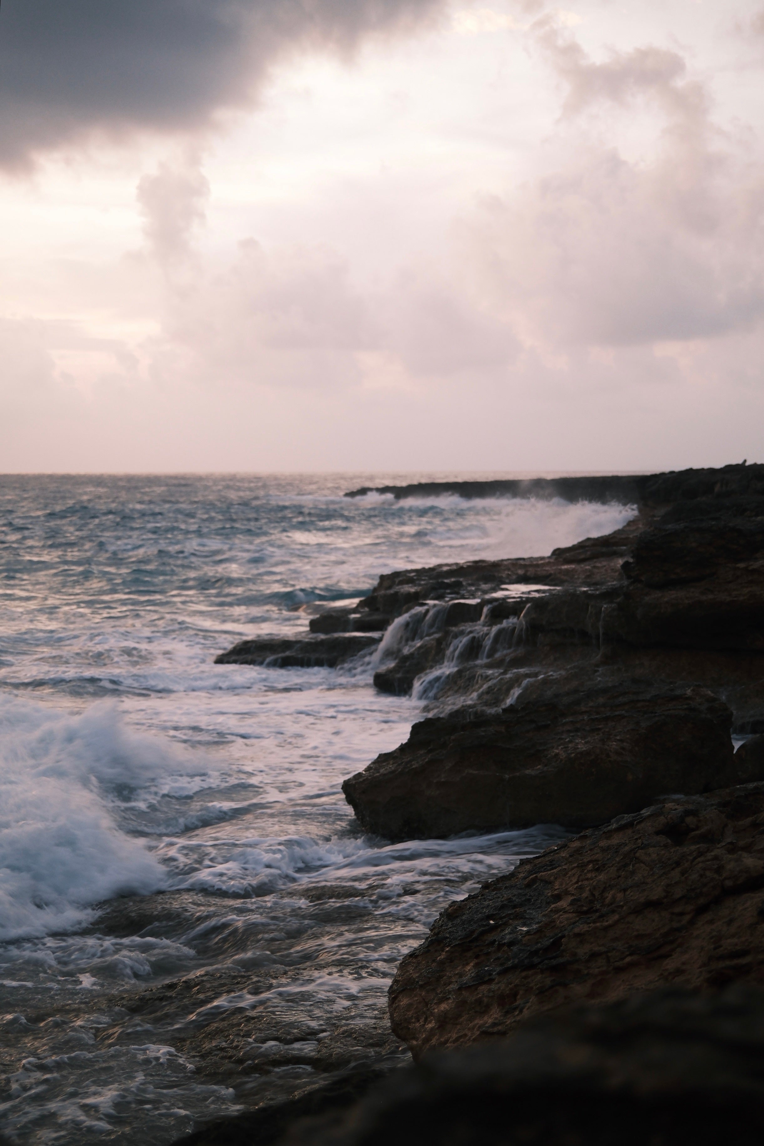 Olas rompiendo contra una costa de rocas oscuras bajo un cielo de nubes rosadas al final del día