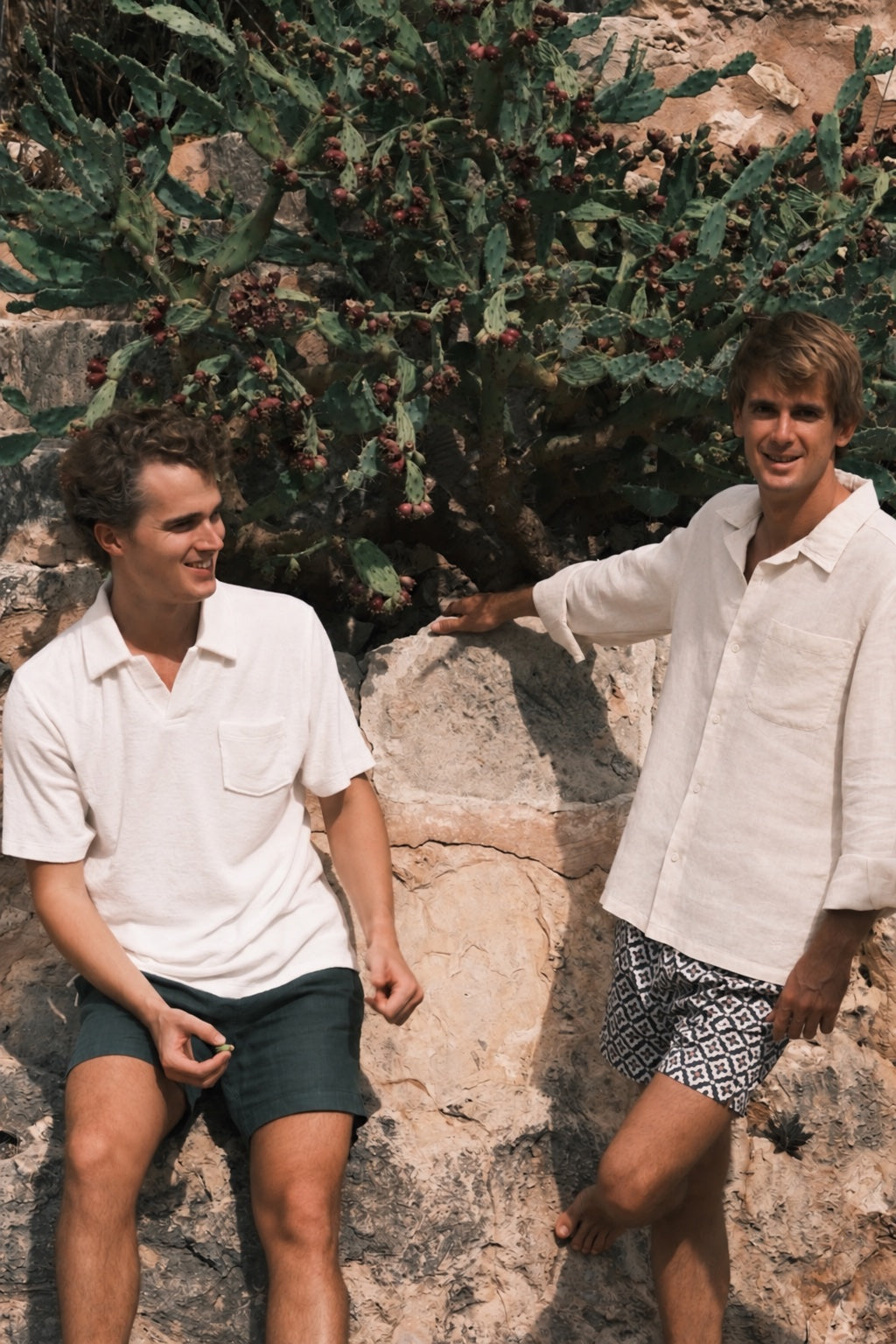 Dos jóvenes apoyados en una roca junto a una chumbera, sonrientes, con un polo blanco y una camisa de lino clara
